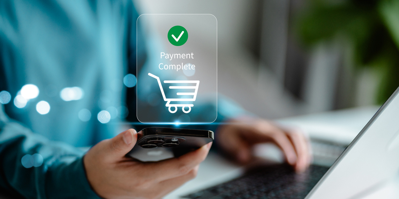 Person holding a phone displaying "Payment Complete" next to a laptop, indicating a successful online transaction.